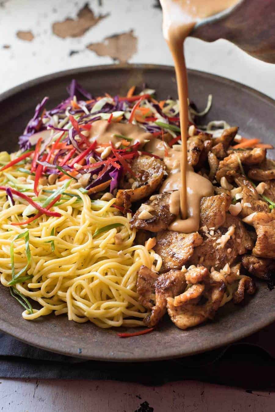 Peanut dressing being poured over Satay Chicken Noodle Salad in a brown bowl.
